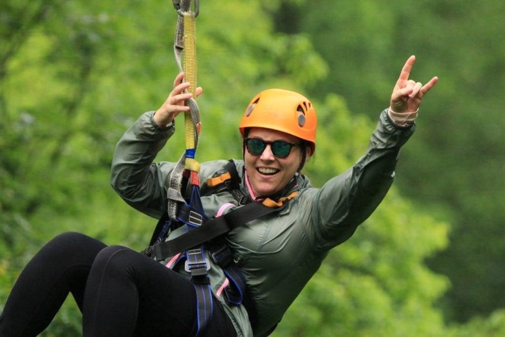 A smiling rider flashing a hand sign while riding the canopy course at Wahoo Ziplines.